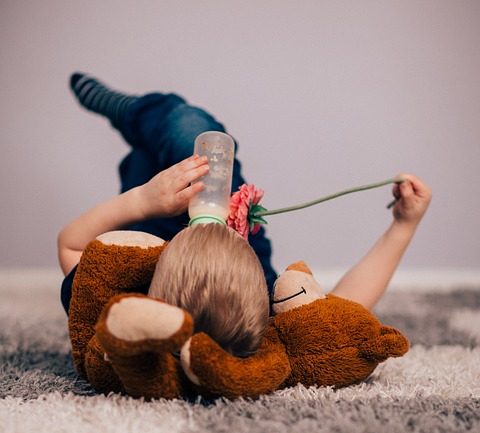 Enfant de dos allongé par terre sur son nounours et buvant un biberon de lait seul